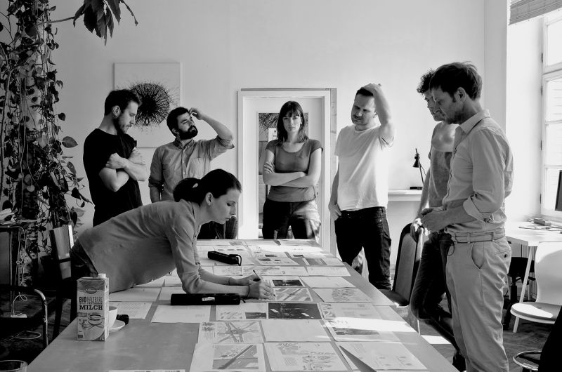 5 Männer und zwei Frauen um einen Tisch versammelt, auf dem viele unterschiedliche Bilder der Radbahn-Planung liegen. Die Personen sehen sehr angestrengt aus und sind am Überlegen. Eine der Frauen beugt sich über den Tisch und zeichnet in eine der Skizzen.