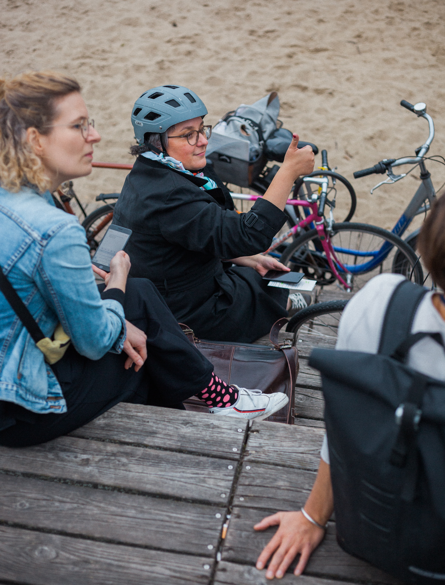 Zwei Frauen sitzen auf Stufen. Das Bild ist von einer darüberliegenden Stufe aufgenommen worden. Die beiden Frauen sitzen wiederum auch auf unterchiedlichen Stufenebenen. Die eine Frau guckt nach recht und hält ein Handy in der Hand. Die darunter sitzende Frau, die einen Fahrradhelm und Brille trägt, schaut ebenfalls nach rechts und zeigt mit ihrem Daumen nach oben und lächelt zustimmend. Es schein als ob rechts im Bilde jemand steht der redet. Die beiden Frauen hören also zu.