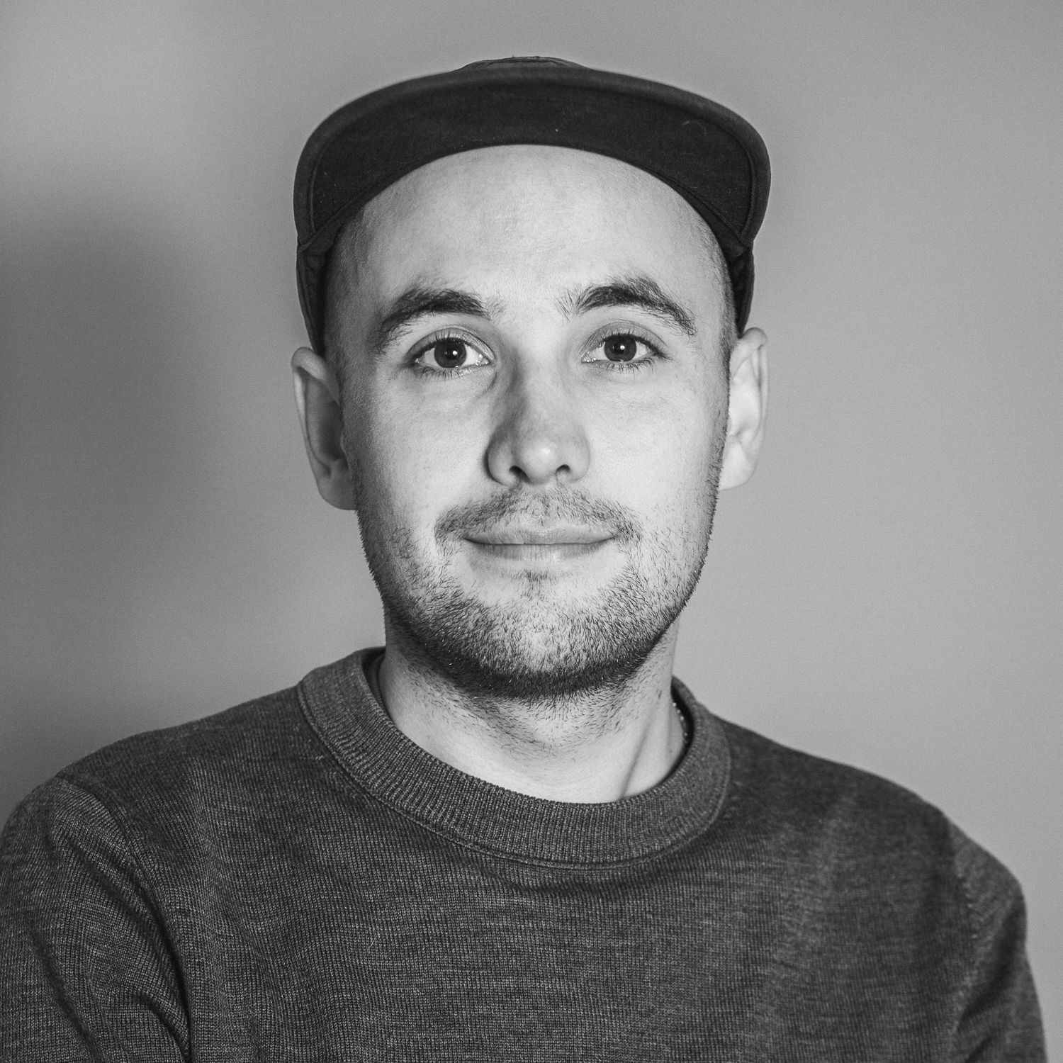 Porträtfotografie eines junger Mannes Anfang 30 mit Fahrradkappe und dunklen Augen und Dreitagebart, der leicht lächelnd frontal in die Kamera guckt.