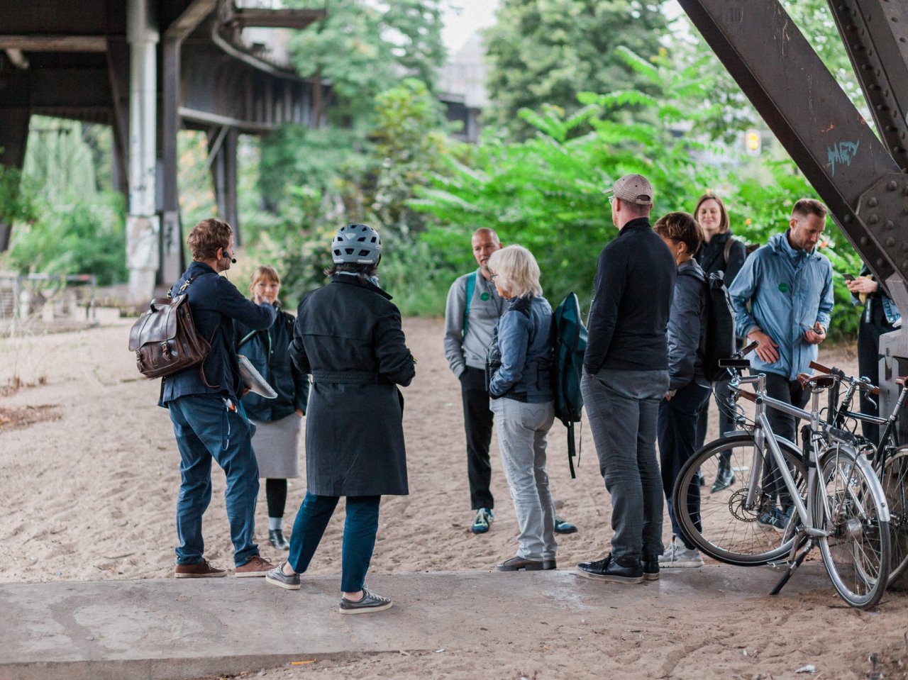 Neun Personen stehen dicht unter dem Viadukt auf Höhe der Möckernbrücke auf sandigem Boden und hören einem Mann zu, der mit Headset da steht und über den Ort erzählt.
