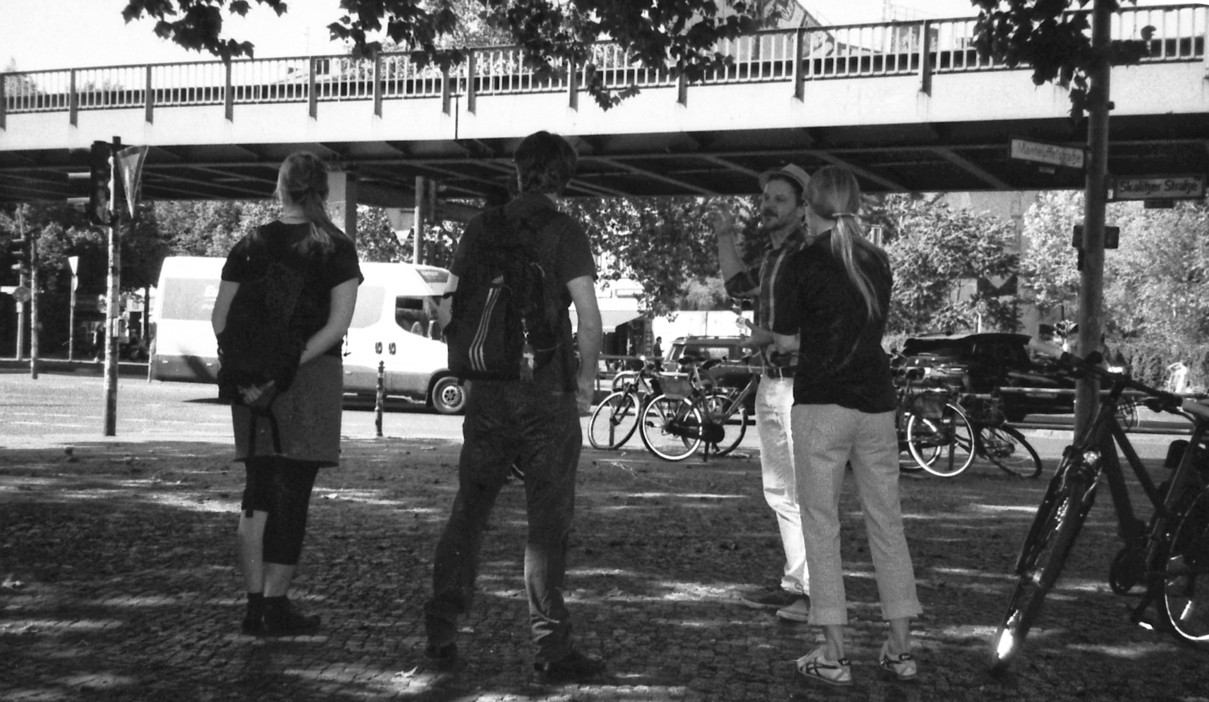 vier personen stehen in einem Kreis und unterhalten sich. Sie stehen unter einem Baum. Hintergrund zeigt die Hochbahn in Berlin Kreuzberg.