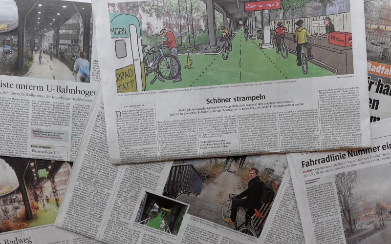 Verschiedene zeitungen wurden auf einen Tischen gelegt und überlappen sich teilweise. Die Zeitungen zeigen deutschsprachige Artikel zur Radbahn. Man liest beispielweise eine Überschrift 