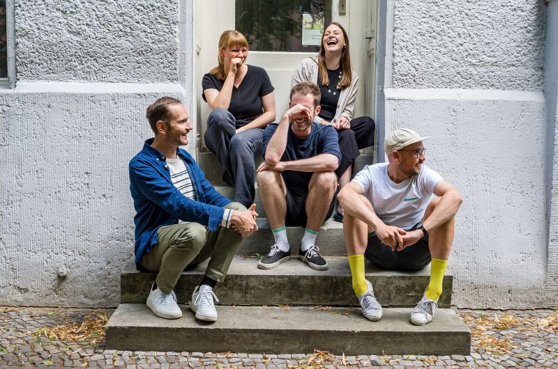 Sechs Personen aus dem Reallabor-Team sitzen vor den Stufen des Büros. In der ersten Reihe drei Männer, in der zweiten Reihe ein Mann, in der obersten Reihe zwei Frauen. Die Personen blödeln rum, gucken nicht in die Kamera und lachen herzlich.