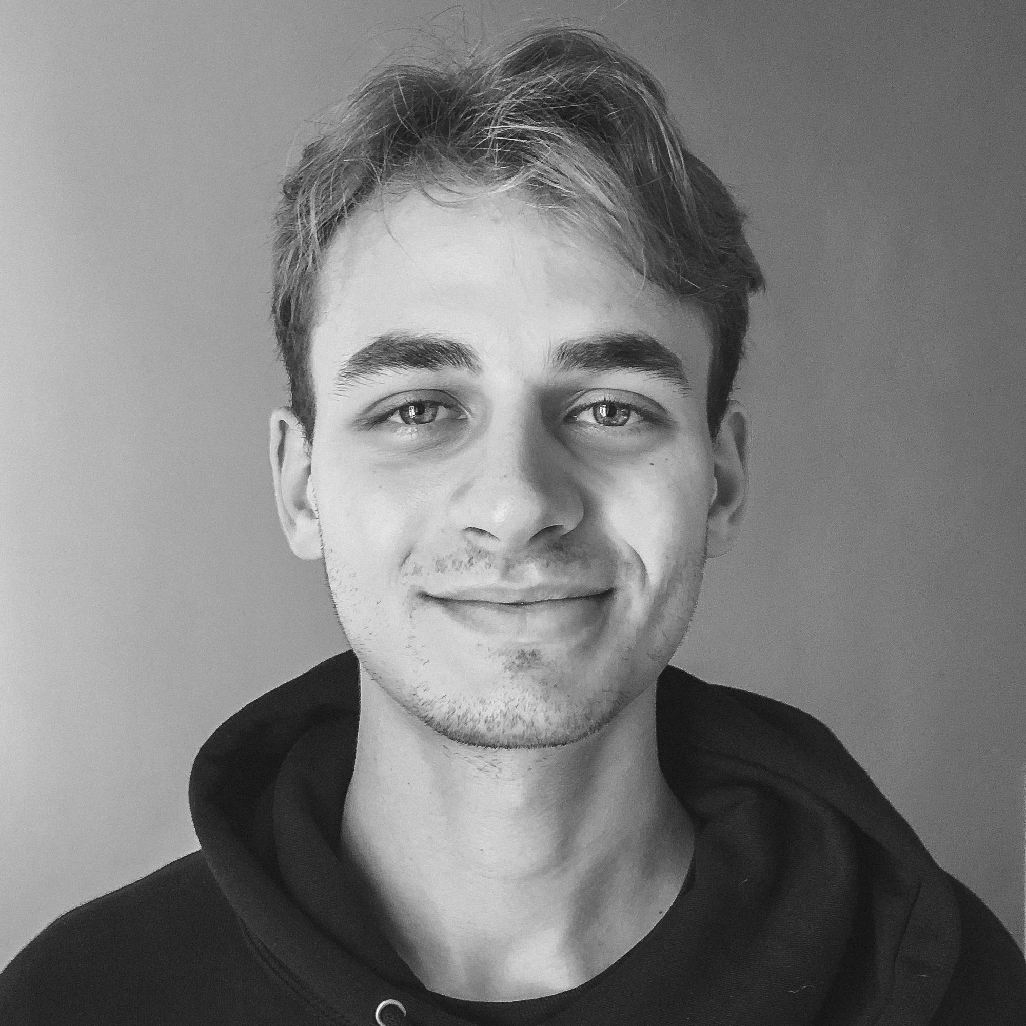 Junger Mann, Anfang bis Mitte 20, Blondes, leicht gewelltes Haar und dominante Augenbrauen,  guckt freundlich lächelnd in die Kamera.