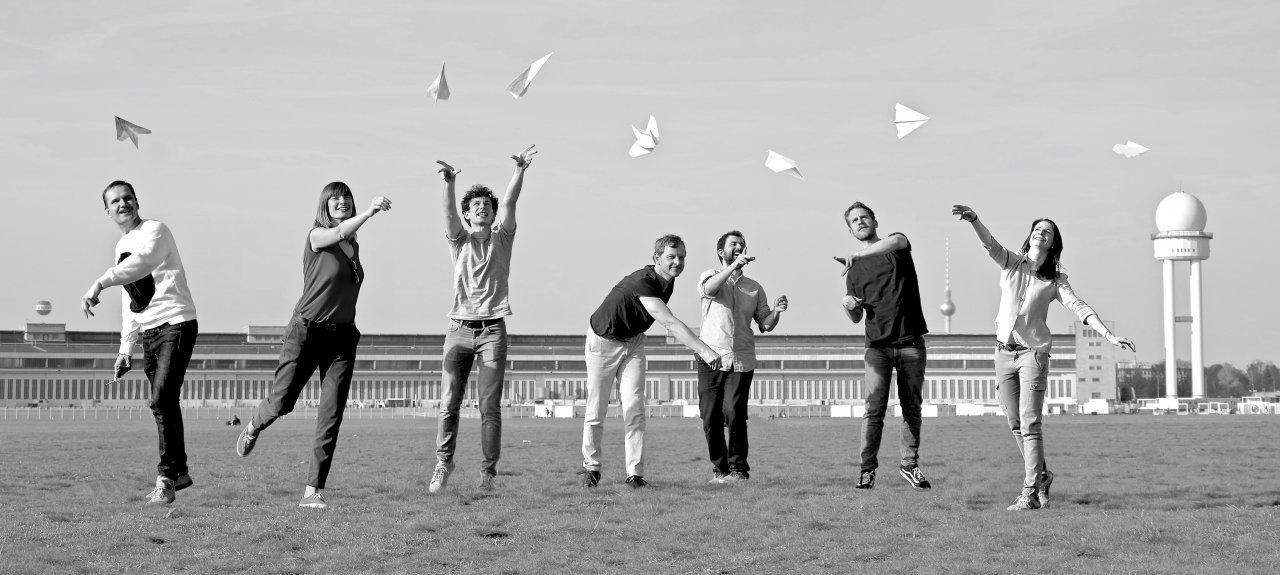Gruppenfoto der Gründer und Gründerinnen von paper planes e.V. Sie stehen auf dem Tempelhofer Feld, im Hintergrund erkennt man das alte Flughafengebäude. Die Personen werfen alle jeweils einen Papiergflieger in die Luft.