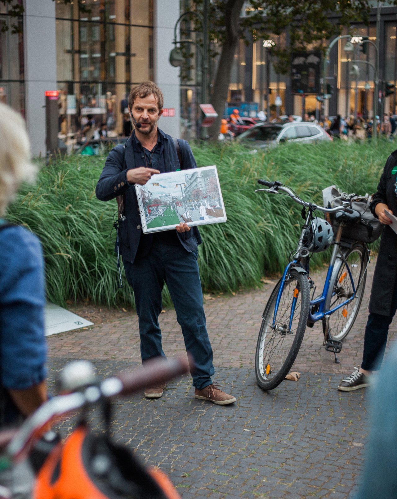 Ein Mann mit Headset hält eine Zeichnung in der Hand. Die Zeichnung zeigt zum Zuschauer. Offensichtlich erklärt er Umstehenden, die nicht im Bilde sind, was auf der Zeichnung abgebildet ist.
