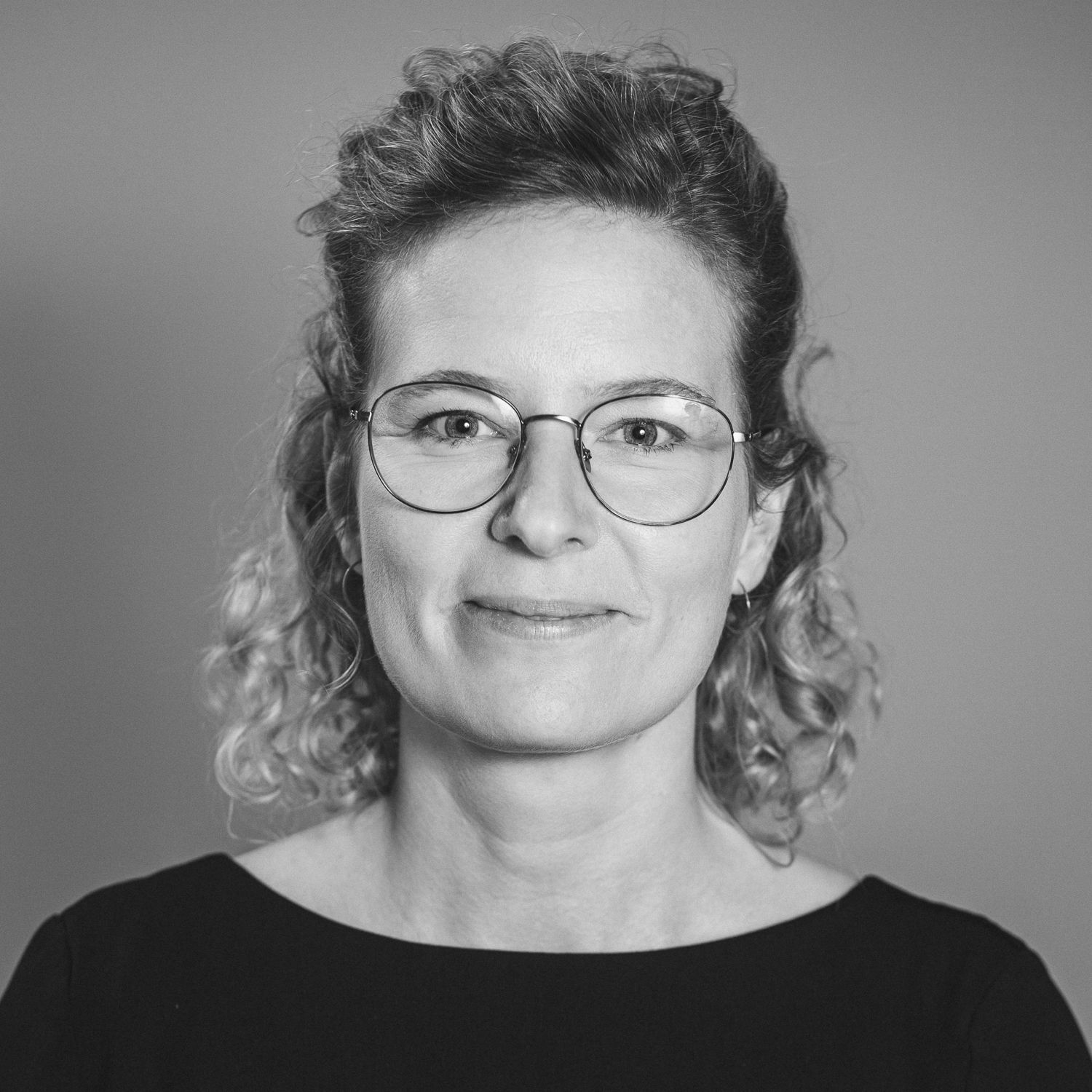 Portraitfotografie einer Frau Anfang 40 mit Brille und geklockten Haaren, die halb zurückgeseteckt sind. Sie lächelt und guckt frontal in die Kamera.