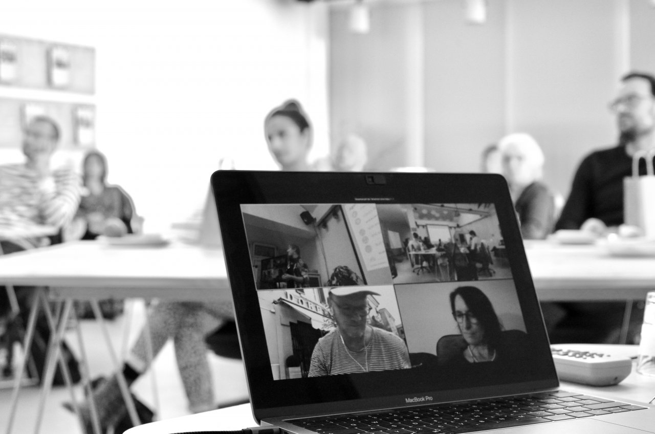 Aufnahme eines Treffen des Projektbeirats. Im Vordergrund ein Bildschirm, der die per Videokonferenz zugeschalteten Teilnehmenden zeigt. Im Hintergrund die im Raum anwesehenden Teilnehmenden.