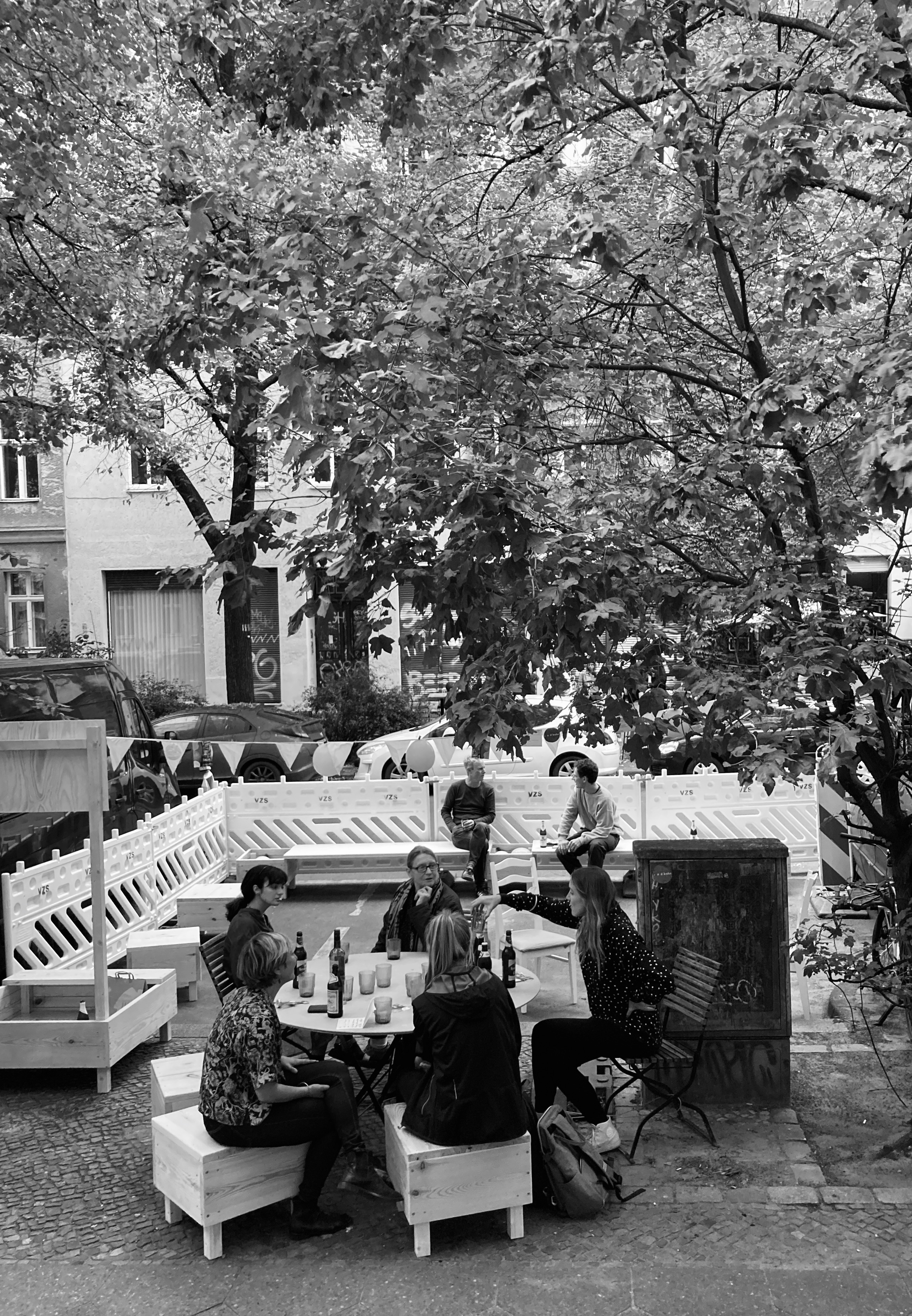 Blick aus dem Büro auf die Forster Strasse, wo im Sommer und Herbst 2021 drei Parkplätze zu einer Terrasse umgewidmet wurden. Eckige Sitzkisten aus Holz und Bänke wurden aufgestellt und der Bereicht mit Baustellenbarken vom Verkehr geschützt. Auf dem Bild sitzen Leute auf diesem Mobiliar um einen Tisch versammelt und unterhalten sich.