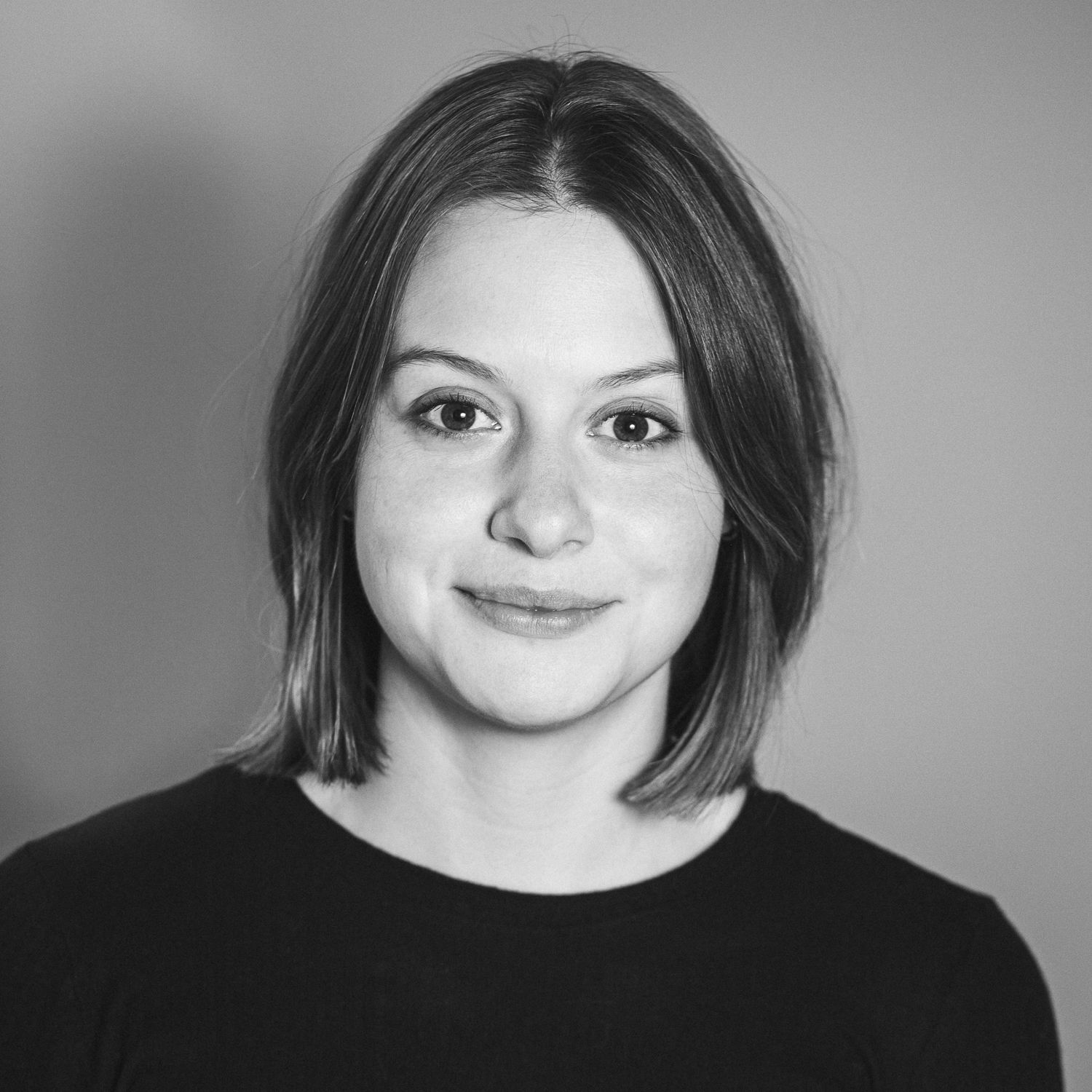 Porträtfogorafie einer jungen Frau Ende 20 mit Schulterlangen braunen Haaren und großen Augen, die mit leicht geneigtem Kopf und lächelnd in die Kamer guckt.