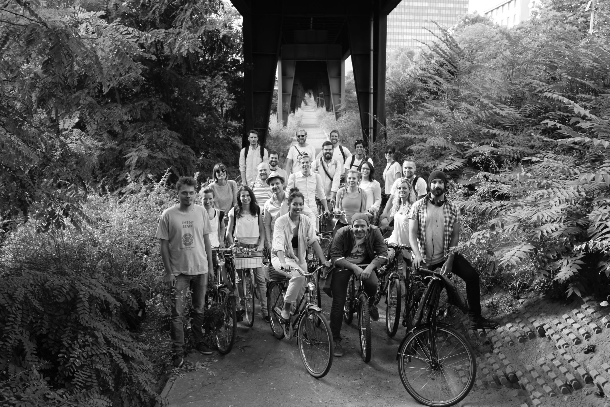 Eine Gruppe von Menschen (ca. 20), dich gedrängt auf Fahrrädern unter dem Hochbahn-Viadukt lächelt als Gruppenfoto in die Kamera. Das Bild wurde leicht erhäht aufgenommen und man sieht im Hintergrund das tunnelartige Viadukt. Links und rechts viel Gebüsch.