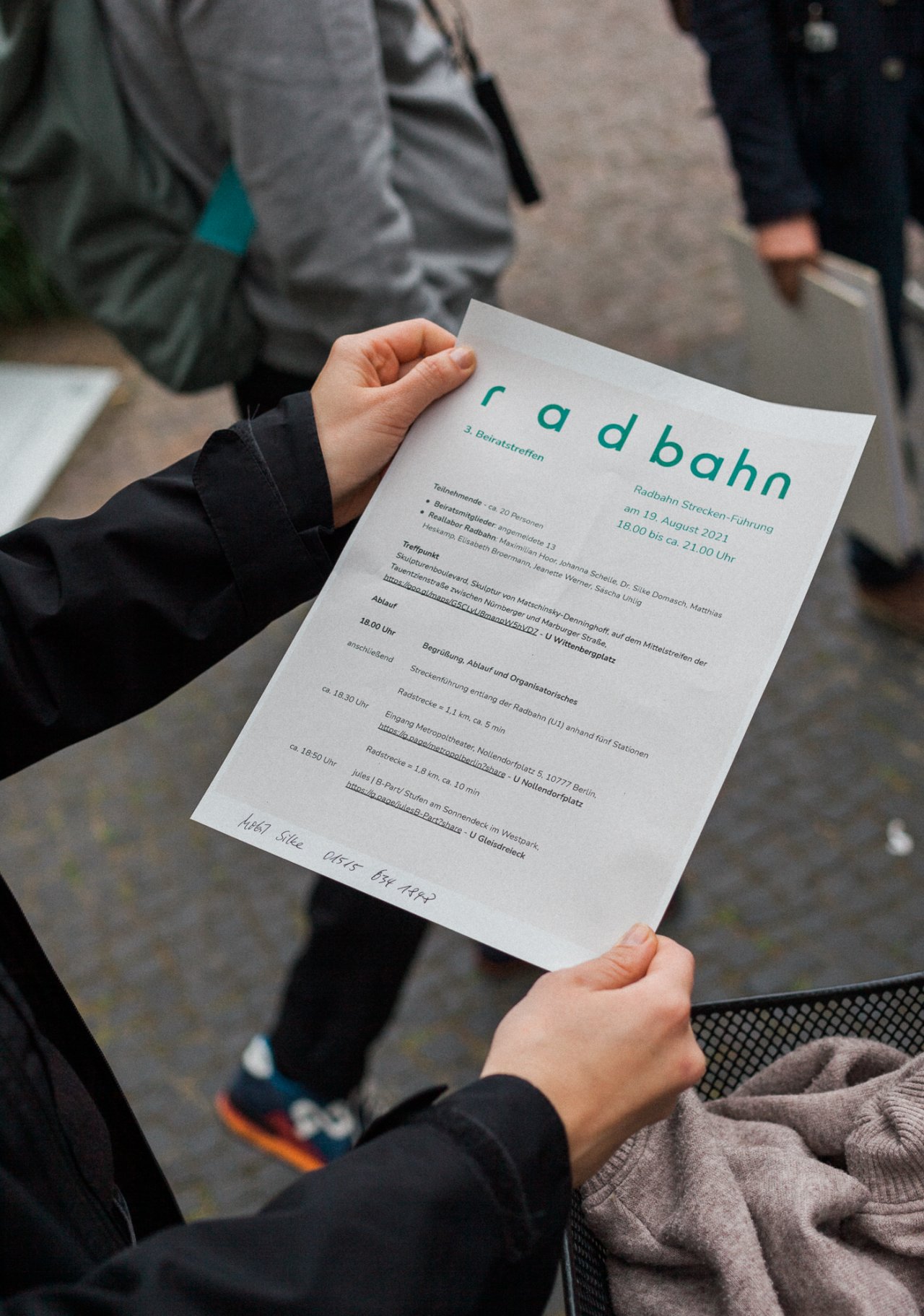 Eine Fotografie, die über die Schulter einer Person fotografiert. Man sieht zwei Hände, die ein DinA4-Blatt halten, auf dem groß das Radbahn-Logo zu sehen ist. Den Rest des Textes kann man nicht gut lesen, aber es handelt sich um eine Agenda oder ein Programm anderer Art.