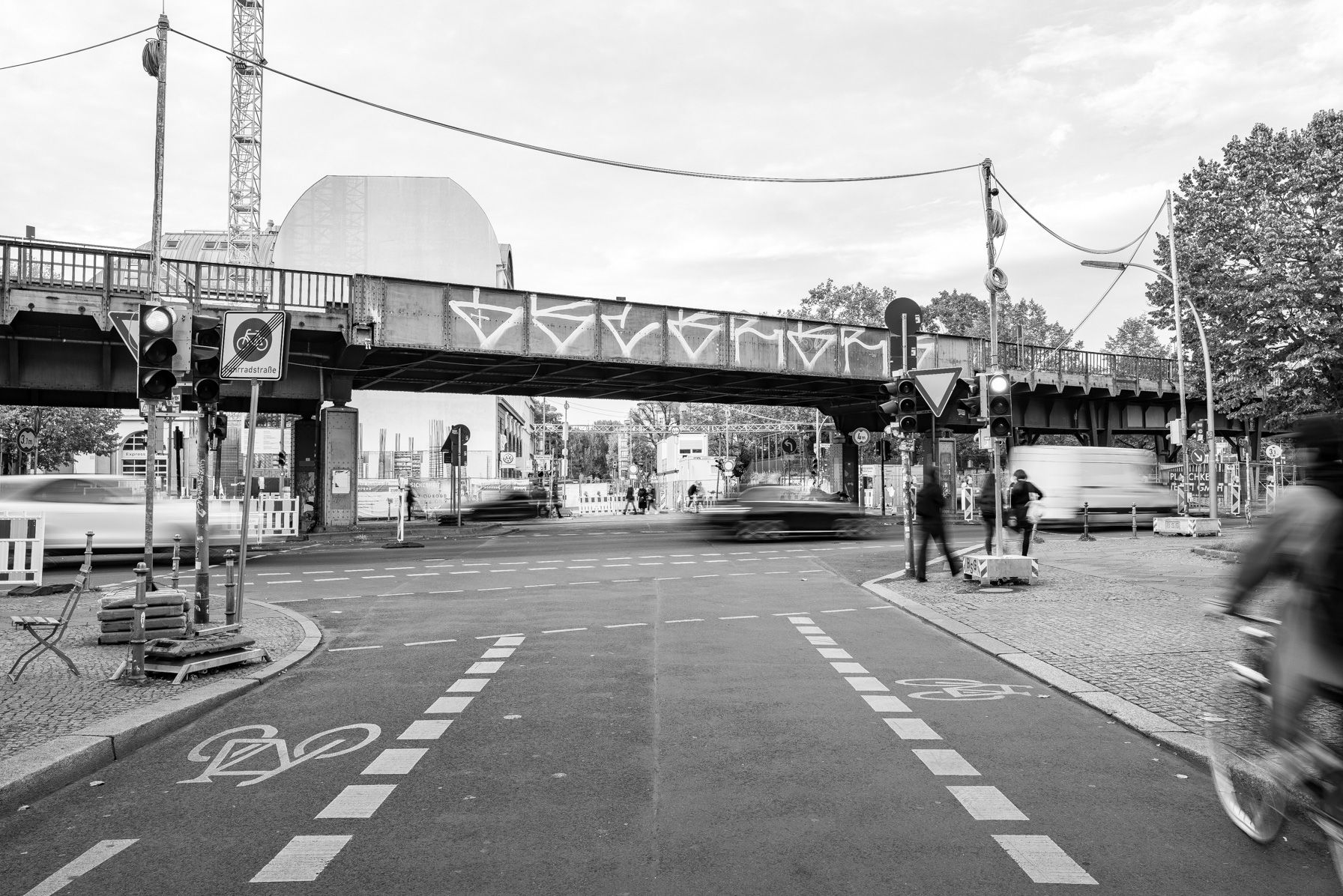 Blick von der Mariannenstraße auf das Hochbahn-Viadukt. Im Vordergrund sieht man an beiden Straßenseiten Radweg-Markerigungen, in der Mitte eine Ampel, die von Fußgängern überquert wird.