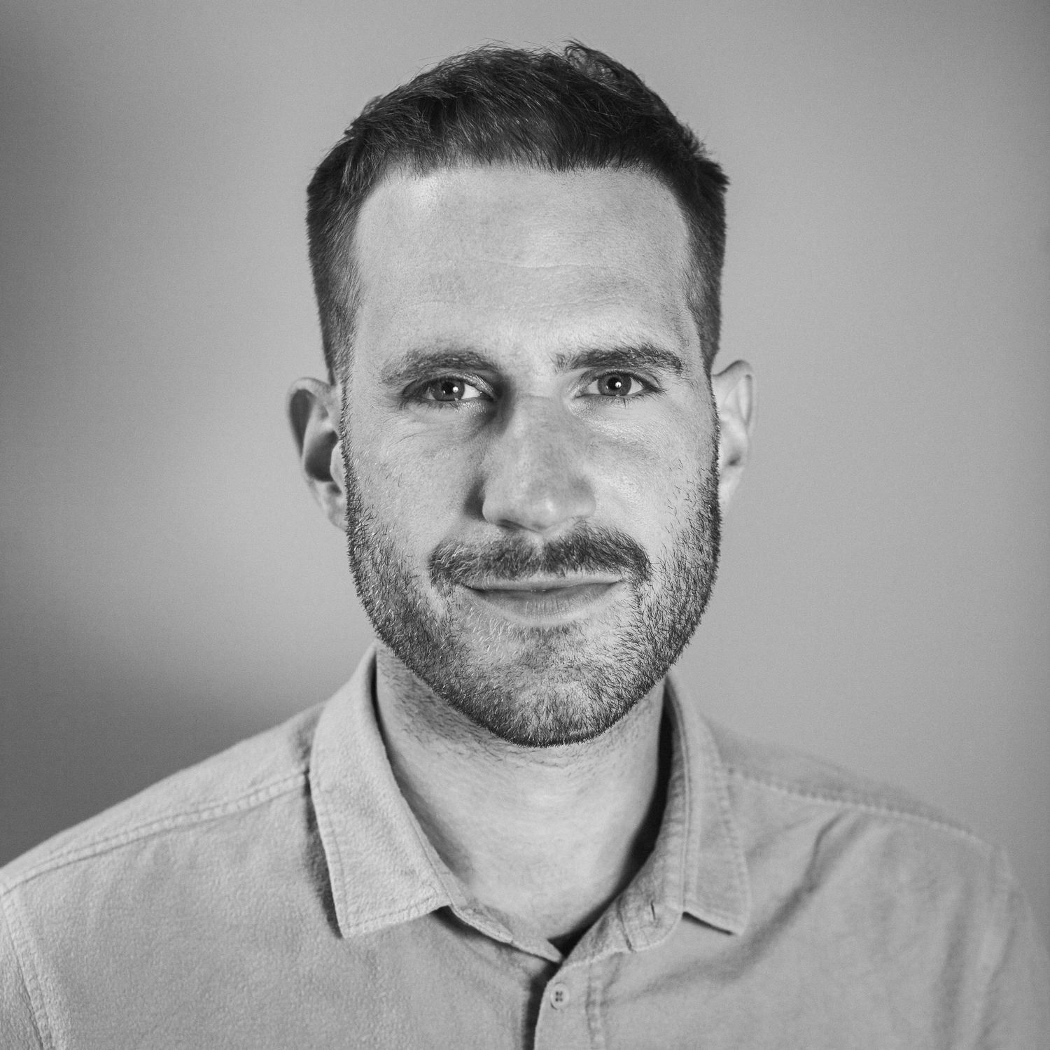 Portritfotografie eines jungen Mannes Mitte 30 mit ordentlichem Kurzhaarschnitt und Dreitagebart, der freundlich in die Kamer blickt.