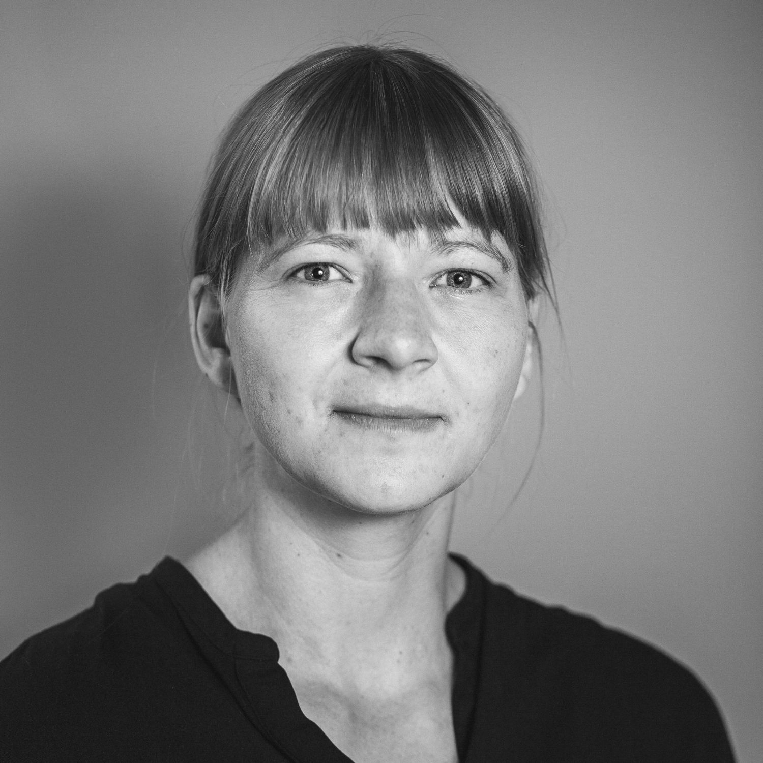 Porträtfotografie einer Frau Anfang 40 mit Pony und blond-brünetten Haar, die zu einem Pferdeschwanz gebunden sind, blickt leicht seitlich, aber dennoch nach vorne, in die Kamera.