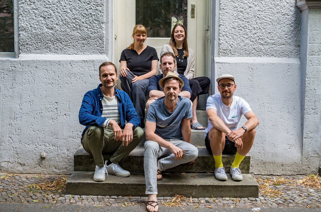 Sechs Personen aus dem Reallabor-Team sitzen vor den Stufen des Büros. In der ersten Reihe drei Männer, in der zweiten Reihe ein Mann, in der obersten Reihe zwei Frauen. Die Personen blicken frontal in die Kamera und lächeln.